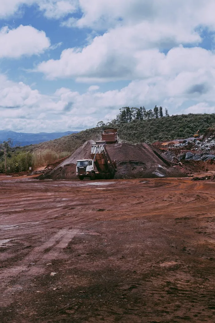 Wide view of a mining site with dump trucks and red dirt, under a cloudy sky.