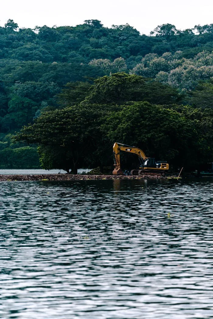 An excavator operates at a construction site by the lake surrounded by dense green forest.