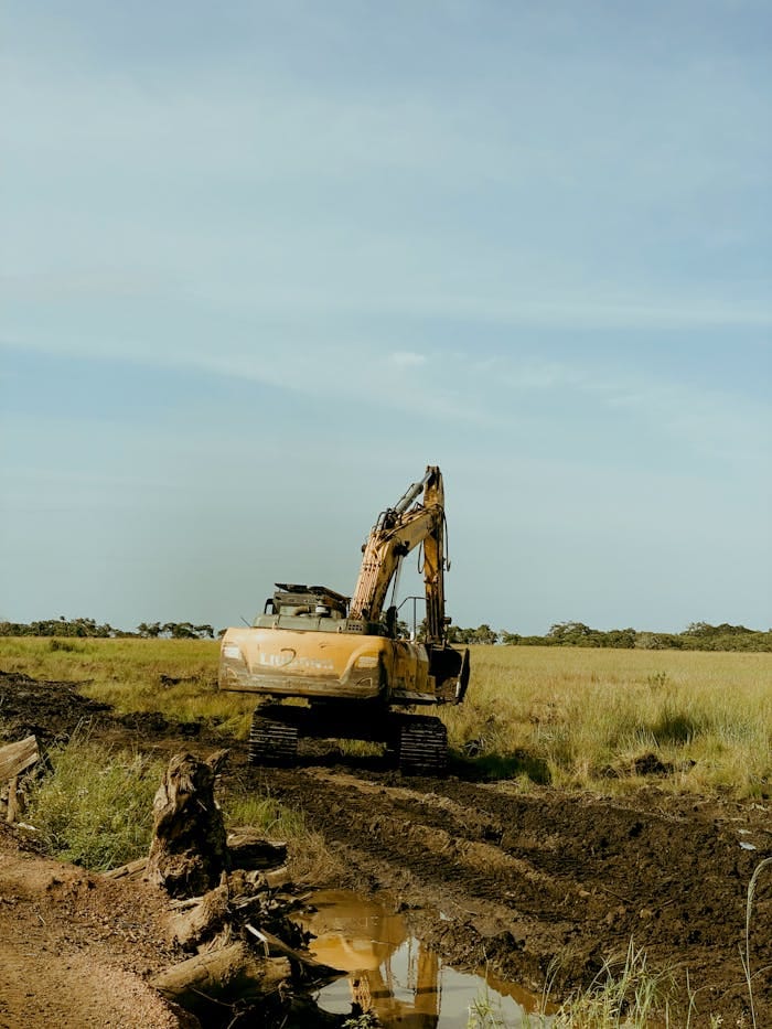 An excavator at work on a rural construction site, surrounded by field and clear sky.