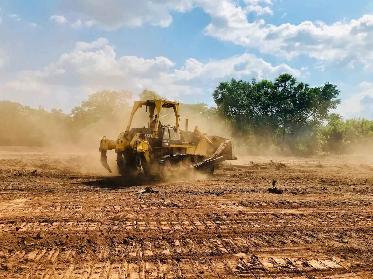 A powerful bulldozer works on a dusty, dirt field under a bright blue sky with cloud cover and trees in the background.
